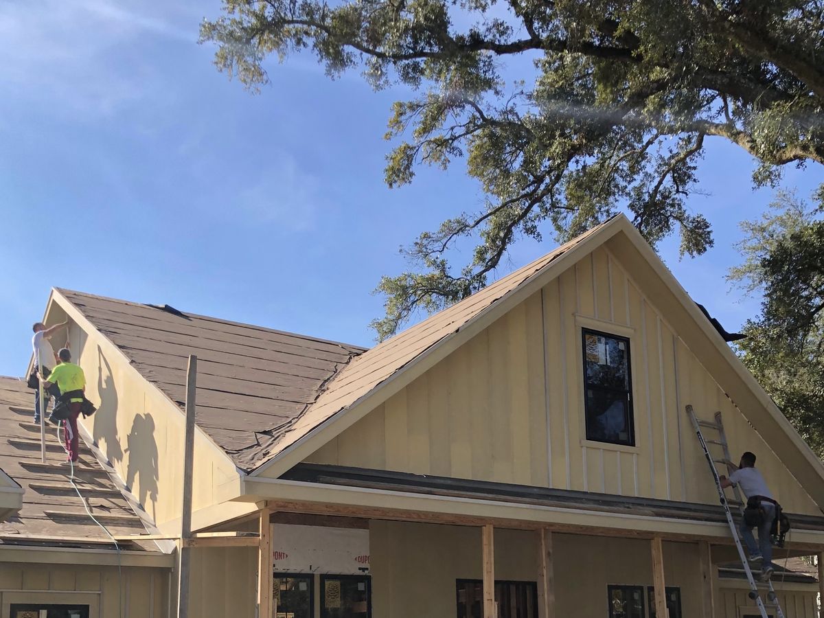 Tan board-and-batten farmhouse with roofers laying brown shingles